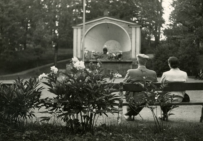 Mustavalkokuvassa kesäisen Pusupuiston eli vanhapuiston lavan edessä kukkaistutusten viereisellä penkillä istuu kaksi henkilöä selin kameraan. Orkesteri on puisella soittolavalla.